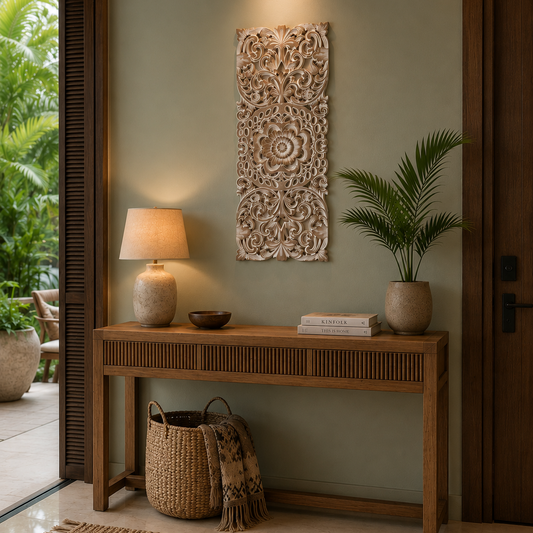 A styled interior entryway featuring a light brown wooden console table against a sage green wall, decorated with a carved beige wall art piece, a table lamp, books, a potted palm, and woven baskets on the floor.